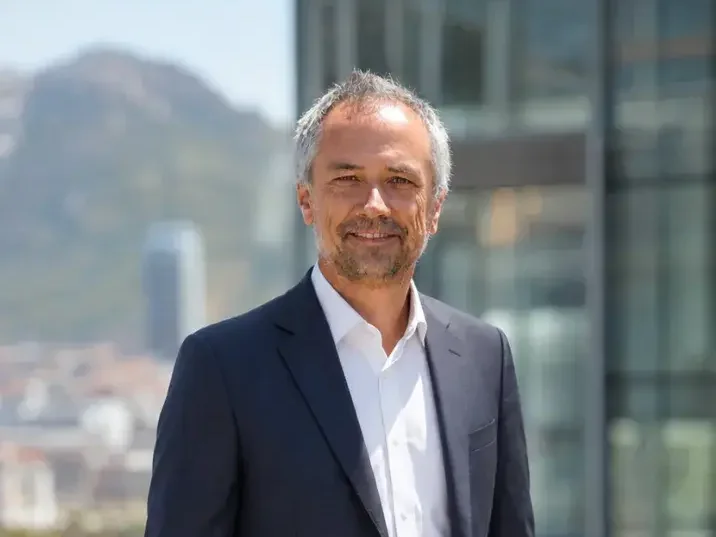 Portrait of Martin Riedi in a navy suit and open-collar white shirt, with a city and mountains behind him